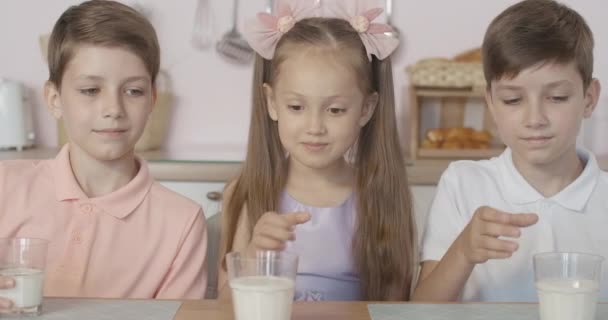 Primer plano de niños felices tintineando vasos con leche y sonriendo. Retrato de niños gemelos caucásicos alegres y niña desayunando con bebida láctea en la mañana . — Vídeo de stock