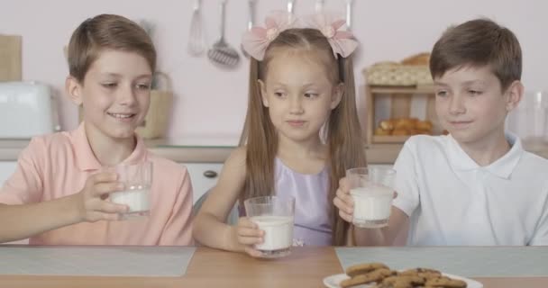 Drie gelukkige blanke kinderen die glazen drinken met melk en smakelijk zuivelproduct. Portret van tevreden kleine tweeling jongens en meisje genieten van het ontbijt in de ochtend thuis. — Stockvideo