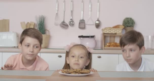 Tres niños alegres asomándose de la mesa, mirando a su alrededor, robando galletas del plato y escondiéndose de nuevo. Retrato de niños y niñas caucásicos positivos comiendo galletas dulces poco saludables en casa . — Vídeo de stock