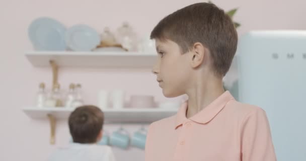 Primer plano de niño caucásico confundido mirando a su alrededor como su hermano gemelo tratando de llegar a platos en la parte superior de la cocina. Retrato de niños lindos descansando en casa. Niños dejados solos en casa sin padres . — Vídeo de stock