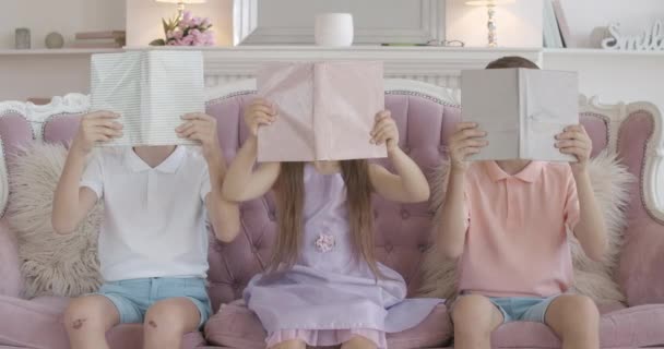Három vicces vidám gyerek ül a kanapén csukott arccal, leteszi a könyveket és mosolyog a kamerába. Portré boldog aranyos kaukázusi ikerfiúkról és lányról, akik otthon olvasnak. Z generáció. — Stock videók