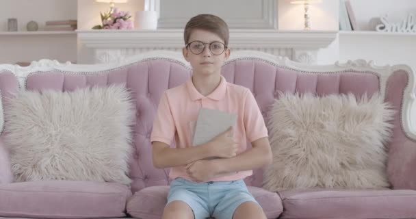 Porträt eines kleinen intelligenten Jungen mit Brille, der mit Buch auf der Couch sitzt und in die Kamera blickt. Kluges kaukasisches Kind posiert drinnen. Nerd brünette schuljunge auf lila sofa mit kissen. — Stockvideo