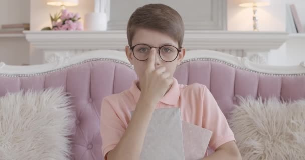 Σοβαρό Καυκάσιο αγόρι που προσαρμόζει γυαλιά και κοιτάζει την κάμερα. Πορτρέτο του γοητευτικό έξυπνο παιδί θέτει με το βιβλίο στον καναπέ στο σπίτι. Έξυπνα παιδιά.. — Αρχείο Βίντεο