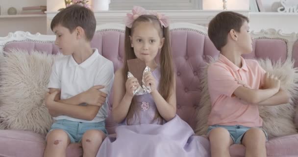 Portré kapzsi kislányról, aki csokit rágcsál, miközben a testvérek mindkét oldalon keresztbe tett kézzel ülnek. A boldogtalan ikerfiúk figyelmen kívül hagyják a nővért, aki nem akar édességet osztani.. — Stock videók
