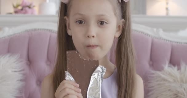 Nahaufnahme eines glücklichen kleinen Mädchens, das zu Hause Schokolade kaut. Zufriedenes Gesicht eines hübschen brünetten kaukasischen Kindes mit braunen Augen, das ungesundes süßes Essen im Haus genießt. Fröhliches Kind isst Dessert. — Stockvideo