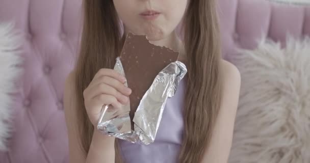 Неузнаваемая маленькая белая девочка, кусающая шоколадку. Крупный план милой брюнетки, наслаждающейся вкусным сладким десертом дома. Дети едят вредную пищу в помещении. — стоковое видео