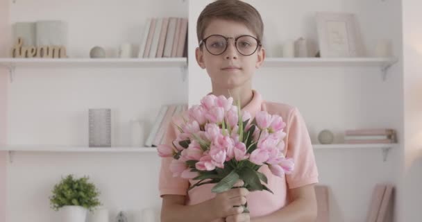 Kocka kisfiú, virágcsokrot nyújtva a kamerának és mosolyogva. Okos fehér srác arcképe szemüvegben, rózsaszín tulipánokkal a könyvespolc hátterében. A gyermek ajándékot ad nyaralásra. — Stock videók