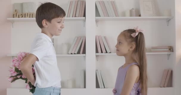 Garçon caucasien donnant bouquet de fleurs à une petite fille timide. Vue de côté portrait de gentleman mignon félicitant joli enfant à l'intérieur. Concept de premier amour, enfance. — Video