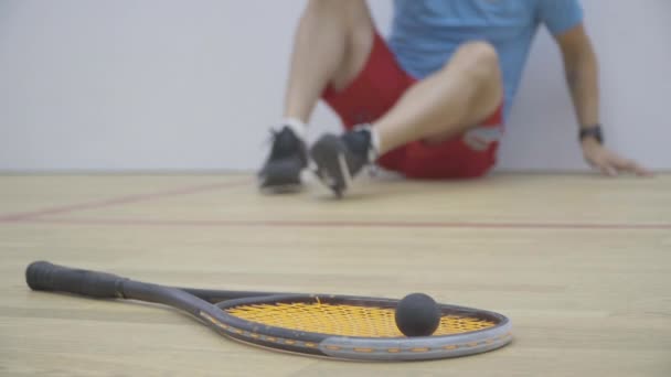 Gros plan de la raquette et du ballon couchés sur le sol dans la salle de gym en tant que sportif caucasien méconnaissable arrivant et prenant de l'équipement. Sportif homme adulte formation dans le sport squash jeu à l'intérieur. — Video