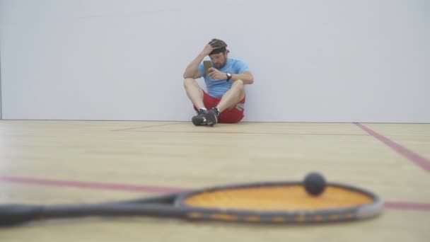 Ευρεία βολή του σοβαρού αθλητή κάθεται στον τοίχο στο γυμναστήριο και χρησιμοποιώντας smartphone. Πορτρέτο της μελαχρινής γενειοφόρος Καυκάσιος άνδρας έχουν διάλειμμα στην εκπαίδευση. Θολή ρακέτα και μπάλα μπροστά. — Αρχείο Βίντεο