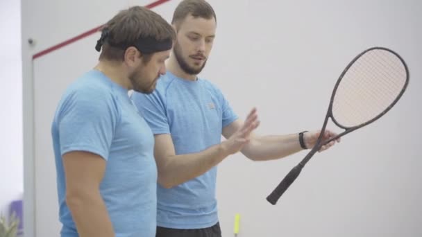 Entraîneur professionnel expliquant comment tenir la raquette de squash à l'homme caucasien mi-adulte. Portrait de jeune entraîneur confiant entrainement brunette barbu gars en salle de gym. Les gens qui jouent au squash sport jeu. — Video