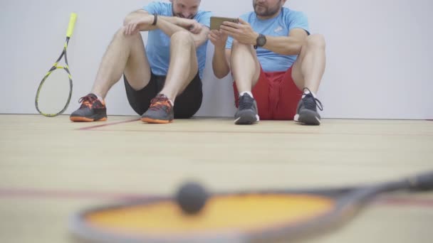 Rack focus change de sportifs positifs à l'aide d'un smartphone et rire de l'arrière-plan pour squash raquette et balle couché à l'avant. Les hommes caucasiens se reposent après la compétition dans la salle de gym. — Video