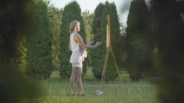 Bred bild av självsäker kvinnlig konstnär som står i solljus i parken och ritar. Sidovy över smal vacker kvinna målning bild vid solnedgången utomhus. Hobby och konst koncept. — Stockvideo