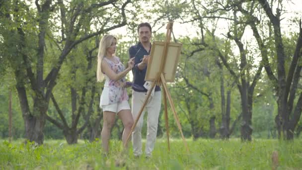 Brede opname van gelukkige blanke schilders die een tekening maken in het zonnige zomerpark. Portret van vrolijke positieve man en vrouw die buiten kunstwerken schilderen. Liefde en verbondenheid. — Stockvideo