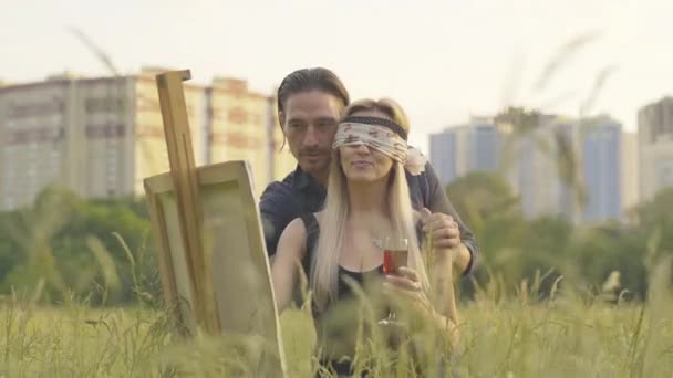 Romantisch Kaukasisch echtpaar schildert landschap bij zonsondergang. Portret van vrolijke vrouw met ogen dicht met sjaal tekening op doek als lachende man die haar helpt te schilderen. Creatieve mensen. — Stockvideo