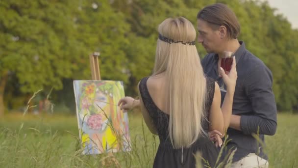 Pohled zezadu na štíhlou blondýnu s vinnou sklovinou obdivující portrétní malbu od pohledného kreativního muže. Šťastný dospělý kavkazský pár stojící venku v parku při západu slunce při pohledu na krásnou kresbu. — Stock video