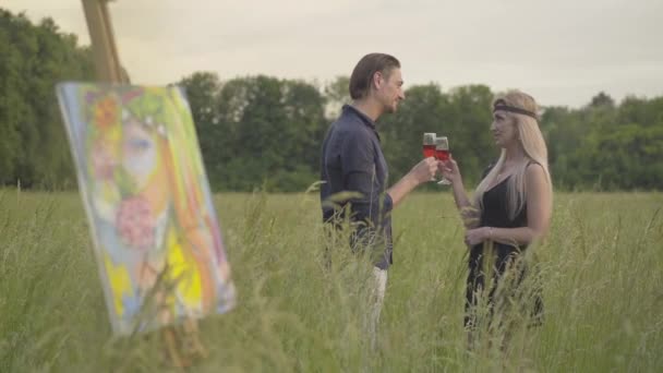 Romantický kreativní pár pije víno a mluví s rozmazaným uměleckým dílem stojícím před ním. Portrét sebevědomého bělocha muž a žena cinkání sklenice po malování venku v letní den. — Stock video