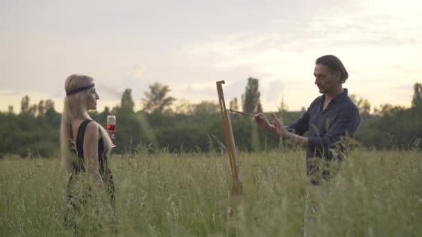 Zijaanzicht van geconcentreerde kunstenaar schilderen prachtige blonde vrouw bij zonsondergang. Professionele mannelijke blanke schilder tekening portret van mooie dame buiten op zomerdag. — Stockvideo