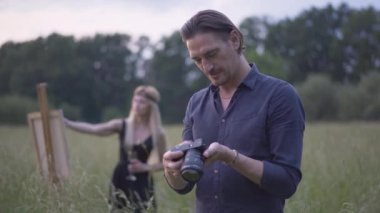Arka planda bulanık, güzel bir ressam resmi gibi fotoğraflara bakan mutlu, yakışıklı fotoğrafçı. Yazın gün batımını dışarıda geçiren yaratıcı kadın ve erkeğin portresi..