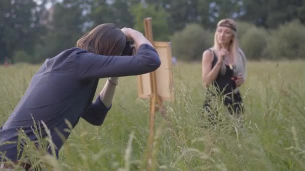 Brunett Kaukasiska mannen tar foto av omtänksam kvinna målning bild på duk. Älskade pojkvän fotografera suddig blond kvinnlig konstnär rita på ängen vid solnedgången utomhus. — Stockvideo