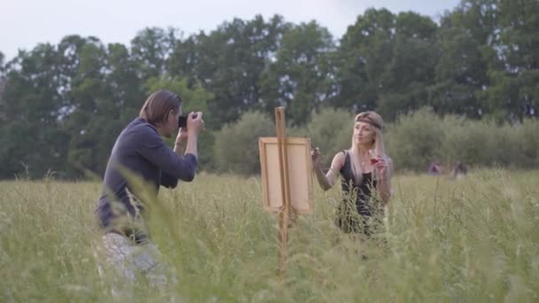 Amplio plano de mujer rubia absorbida con pintura de vidrio sobre lienzo al atardecer como hombre creativo tomando sus fotos. Pareja confiada de artistas disfrutando de la noche de verano al aire libre en el prado. — Vídeo de stock