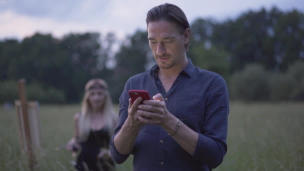 Portrait of brunette Caucasian man using smartphone as blond smiling woman coming from the background. Happy adult couple resting on meadow at dusk. — Stock Video