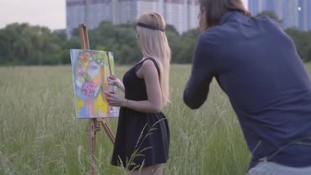 Soustředěná zamyšlená žena kreslí malbu jako muž fotografuje proces tvorby. Pohled zezadu na muže fotografujícího umělkyni při tvorbě uměleckého díla. — Stock video