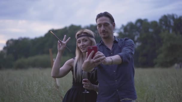 Középső lövés mosolygó kaukázusi kreatív pár, akik szelfit készítenek alkonyatkor a nyári réten. Portré boldog felnőtt férfi és nő szórakozás pihenés szabadban. — Stock videók