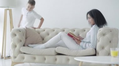 Gülümseyen Afro-Amerikan kadın kanepede uzanıyor ve etrafta koşuşturan neşeli çocuk olarak kitap okuyor. Mutlu aile hafta sonları. Neşeli anne ve oğul evde dinleniyor..