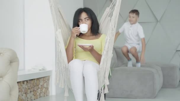 Calm African American woman drinking tea or coffee as active son jumping on bed at the background. Carefree mother ignoring disobedience of child at home. Sedatives abuse against stress. — Stock Video