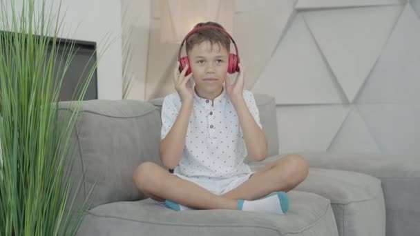 陽気なアフリカ系アメリカ人の少年がヘッドフォンをして音楽に移動します。自宅で趣味を楽しむ楽しいかわいい子供の肖像画。幸せな子供はソファの上に屋内に座って楽しんでいます. — ストック動画