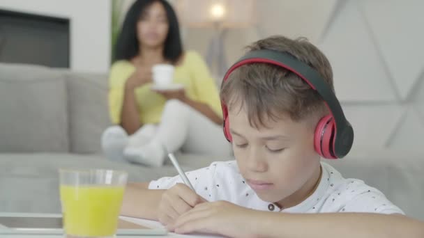 Gros plan du petit garçon afro-américain dans un casque faisant ses devoirs en tant que mère floue buvant du thé ou du café en arrière-plan. Enfant sérieux se déplaçant tête à la musique et l'écriture dans le cahier d'exercices. — Video