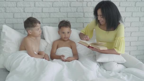 Alegre madre afroamericana leyendo libro para hijos acostados en la cama. Retrato de una joven feliz acostando niños por la noche. Familia feliz descansando en el interior. — Vídeo de stock