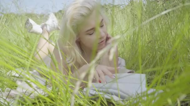 Absorbida mujer de talla grande acostada en hierba verde alta y libro de lectura. Retrato de una joven mujer caucásica con sobrepeso en ropa interior blanca y zapatillas de deporte descansando en un día soleado en el prado. — Vídeos de Stock