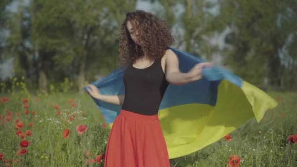 Mooie slanke Oekraïense vrouw spinnen met nationale vlag op zomerveld van papavers. Portret van charmante jonge Kaukasische dame met symbool van de staat genieten van zonnige dag op de zomerweide. — Stockvideo