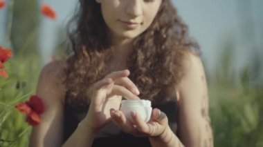 Genç ve güzel bir kadın yüz kremi kavanozunu açıyor ve nemlendirici sürüyor. Güneşli yaz gününde haşhaş tarlasında doğal organik kozmetik kullanan beyaz bir kadın. Cilt bakımı ve güzellik.