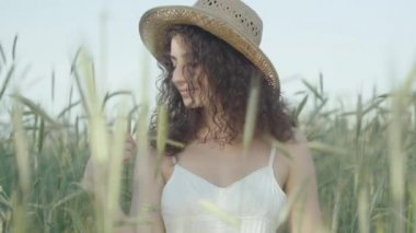 Beyaz elbiseli, hasır şapkalı, bedene çavdar dalıyla dokunan şehvetli genç bir kadın. Açık havada güneşli bir günün tadını çıkaran kıvırcık saçlı ve kahverengi gözlü baştan çıkarıcı esmer kadın portresi..