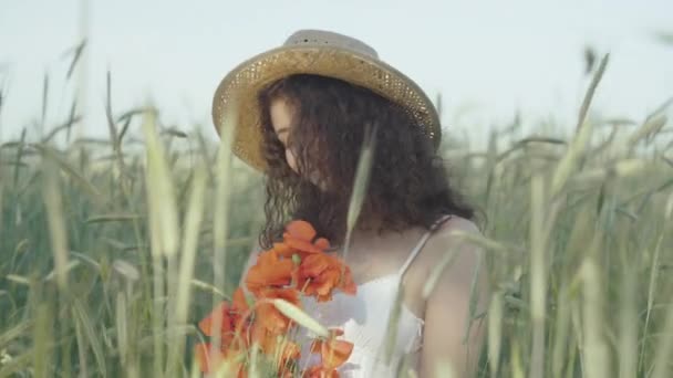 Retrato de mujer joven feliz en sombrero de paja oliendo ramo de amapolas rojas al aire libre y sonriendo. Hermosa encantadora dama caucásica disfrutando de día soleado en el campo de verano. — Vídeo de stock