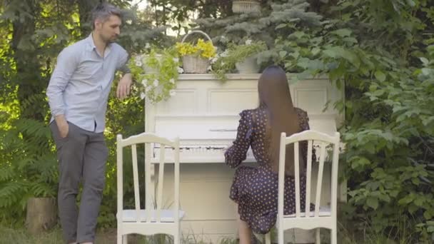 Široký záběr šťastného muže středního věku, který stojí vedle klavíru a poslouchá brunetku, jak hraje hudbu. Portrét pozitivních kreativních bělošských párů trávících víkendy společně venku. — Stock video