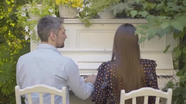 Back view of Caucasian romantic couple playing piano outdoors on summer day. Camera approaches to smiling loving man and woman enjoying sunny day outdoors. Dating concept. — Stock Video