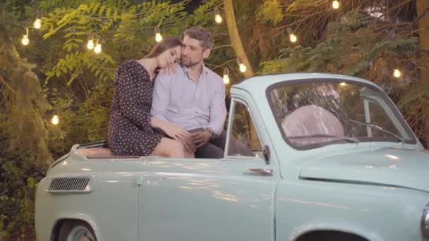 Aparat zbliża się do zrelaksowanej kochającej się pary siedzącej w samochodzie retro i rozmawiającej. Portret szczęśliwego białego mężczyzny i kobiety spotykających się w letni wieczór na świeżym powietrzu. Pojęcie romantyzmu. — Wideo stockowe