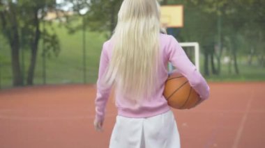 Kamera, sarışın genç bir bayanı dışarıda basketbol sahasında yürürken izliyor. Güneşli yaz gününde, spor sahasında top gezintisi yapan zayıf beyaz kadın. Sağlıklı yaşam tarzı kavramı.