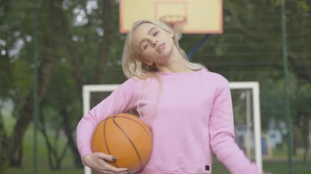 Câmera se aproxima para jovem desportista confiante posando com bola de basquete na quadra ao ar livre. Retrato de bela mulher loira branca magro em pé no chão de esportes sorrindo e olhando para a câmera. — Vídeo de Stock
