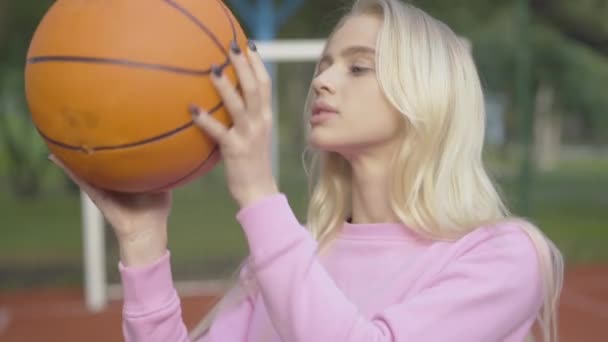 Close-up retrato de jovem bela mulher girando bola de esportes ao ar livre e olhando para a câmera. Desportista loira caucasiana confiante posando na quadra de basquete ao ar livre no dia de verão. — Vídeo de Stock