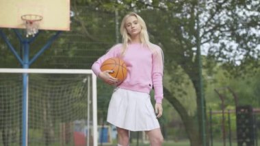 Genç ve güzel bir kadın basketbol sahasında spor topuyla durup gülümsüyor. Kamera, güneşli yaz gününde açık havada poz veren kendine güvenen beyaz kadın oyuncunun etrafında dönüyor..