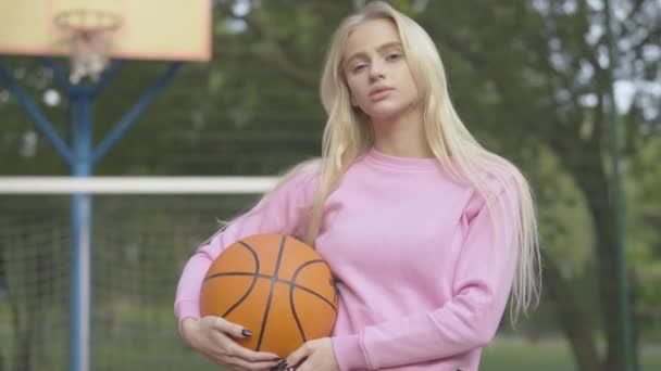 Tiro médio de jovem jogador de basquete confiante posando com bola ao ar livre. Retrato de mulher loira caucasiana sorridente feliz em pé na quadra no dia de verão. Desportista encantadora no parque infantil. — Vídeo de Stock