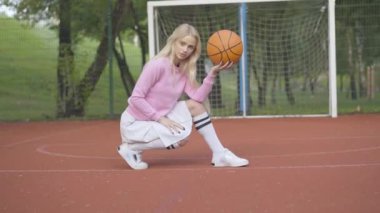 Genç duygusal basketbolcu sahada top ile oturuyor. Yazın oyun parkında spor malzemeleriyle poz veren muhteşem beyaz tenli sarışın kadına kamera yaklaşıyor..