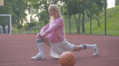 Basketbol maçından önce sahada ısınan kendine güvenen beyaz bir kadının yan görüntüsü. Sarışın güzel oyuncunun portresi. Bacaklarını esnetiyor. Top önde yatıyor..