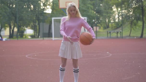 Wanita pirang cantik menggerakan bola basket di tangan dan melihat ke kamera. Wanita olahragawan muda Kaukasia yang percaya diri berpose di luar ruangan di lapangan olahraga. Portrait of beautiful cheerleader. — Stok Video