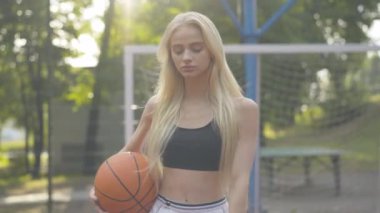 Basketbol sahasında gün ışığında yürüyen zayıf genç bir kadın. Açık havada spor giyimli, kendine güvenen sarışın beyaz bir oyuncunun portresi. Çekici bir kadın, güneş ışığında top oynuyor..